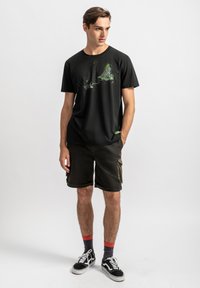 Zwarte t-shirt met een groene grafische ontwerp, gecombineerd met donkere cargo shorts en zwarte sneakers. Sokken zijn marineblauw met rode accenten.