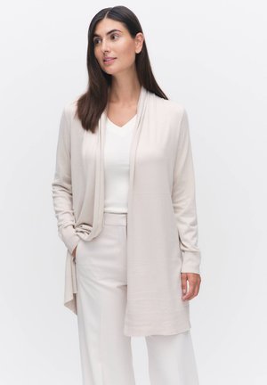 Ljusbeige cardigan med öppen framsidal, tillverkad av mjukt material, har långa ärmar och en avslappnad passform, kombinerad med en vit topp och matchande byxor.