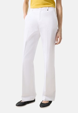 Person iført hvide skræddersyede bukser, gul top stukket ind og sorte flade sko, stående med hænderne i lommerne mod en ensfarvet baggrund.