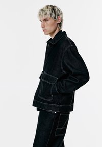 Ung person med lyst hår iført sort denimjakke og bukser med hvid syning, der står i profil mod en hvid baggrund.