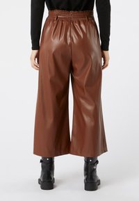 Persona che indossa pantaloni in finta pelle marrone a vita alta e gamba larga, con stivaletti neri, in piedi contro uno sfondo bianco.