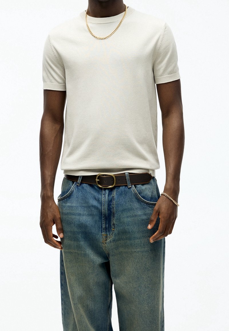 Homme portant une chemise beige à manches courtes, un jean bleu avec une ceinture noire, un collier en chaîne d'or et un bracelet en or sur fond blanc.