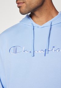 Helles blaue Kapuzenjacke aus strukturiertem Material, mit einem auffälligen gestickten "Champion"-Logo in passendem Farbton und Kordeln an der Kapuze.