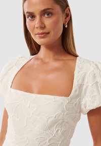 Weißes Kleid mit quadratischem Ausschnitt und Puffärmeln, verziert mit strukturierten floralen Stickereien und seidigem Finish. Goldene Creolen ergänzen den Look.