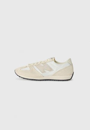 Baskets basses beige et blanches avec des accents en daim, semelle noire à crampons et lacets, vues de profil sur un fond uni.