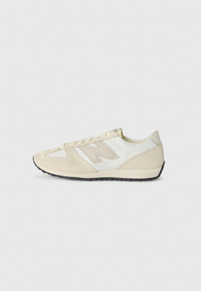 Baskets basses beige et blanches avec des accents en daim, semelle noire à crampons et lacets, vues de profil sur un fond uni.