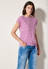 Haut rose à manches courtes avec un motif de feuilles, en tissu léger, encolure ronde et détail à cordon au niveau de l'ourlet, associé à un jean bleu.