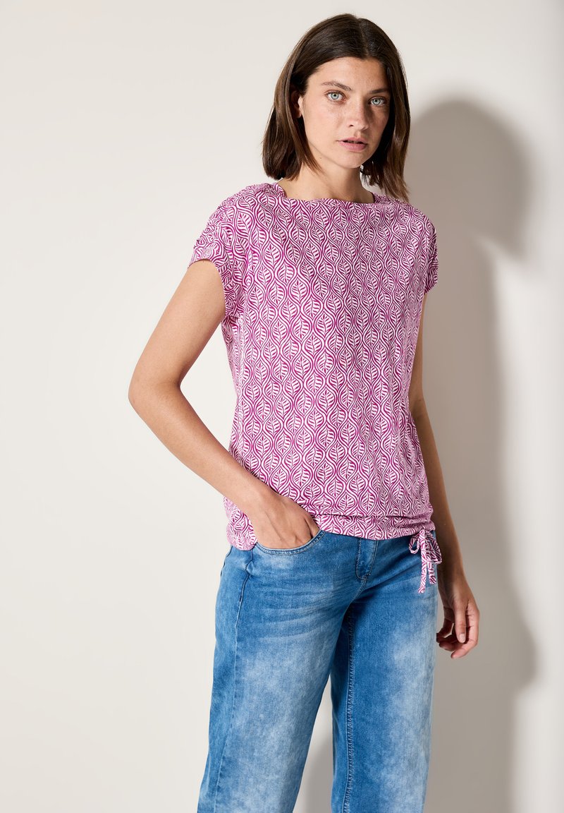Haut rose à manches courtes avec un motif de feuilles, en tissu léger, encolure ronde et détail à cordon au niveau de l'ourlet, associé à un jean bleu.