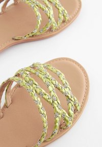 Platta sandaler med en beige sula och flätade gröna remmar med silverdekorationer. Slät textur och öppen design.