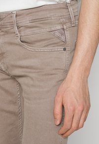 Beiga denimjeans med smal passform, med en sydd ficka och en "REPLAY" logotagg. Hand vilande på sidan, exponerad tygtextur.