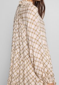 Light beige blouse with a grid pattern in gold and blue, featuring gathered details at the neck and cuffs for a flowing design.