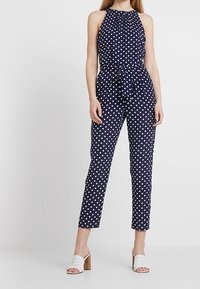 Granatowy kombinezon w kropki z dekoltem na halter, przewiązanym w talii i zwężanymi nogawkami. Połączony z białymi sandałami na obcasie typu block heel z odkrytymi palcami.