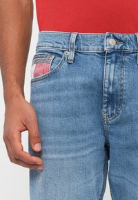 Ανοιχτό μπλε τζιν παντελόνι με ίσια γραμμή, με πολύχρωμο πατς τσέπη και ασημένιο κουμπί στη ζώνη.