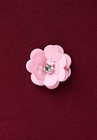 Roze paillettenbloem met een centrale heldere rhinestone, vastgepind op een donkerrode gebreide stofachtergrond.