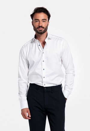 Witte button-up shirt met contrasterende donkere knoopdetails, gecombineerd met donkere broeken, met een klassieke kraag en lange mouwen.