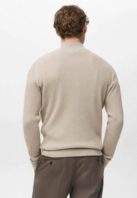 Beiger, gerippter Pullover mit langen Ärmeln und rundem Ausschnitt, strukturierter Stoff, getragen über dunkelbraunen Hosen. Die Ansicht zeigt die Rückseite und Schultermotive.