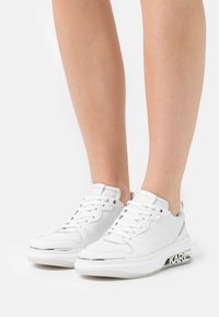 Białe sneakersy na sznurówki z grubymi podeszwami z brandingiem "KARL", noszone przez osobę stojącą na białym tle.