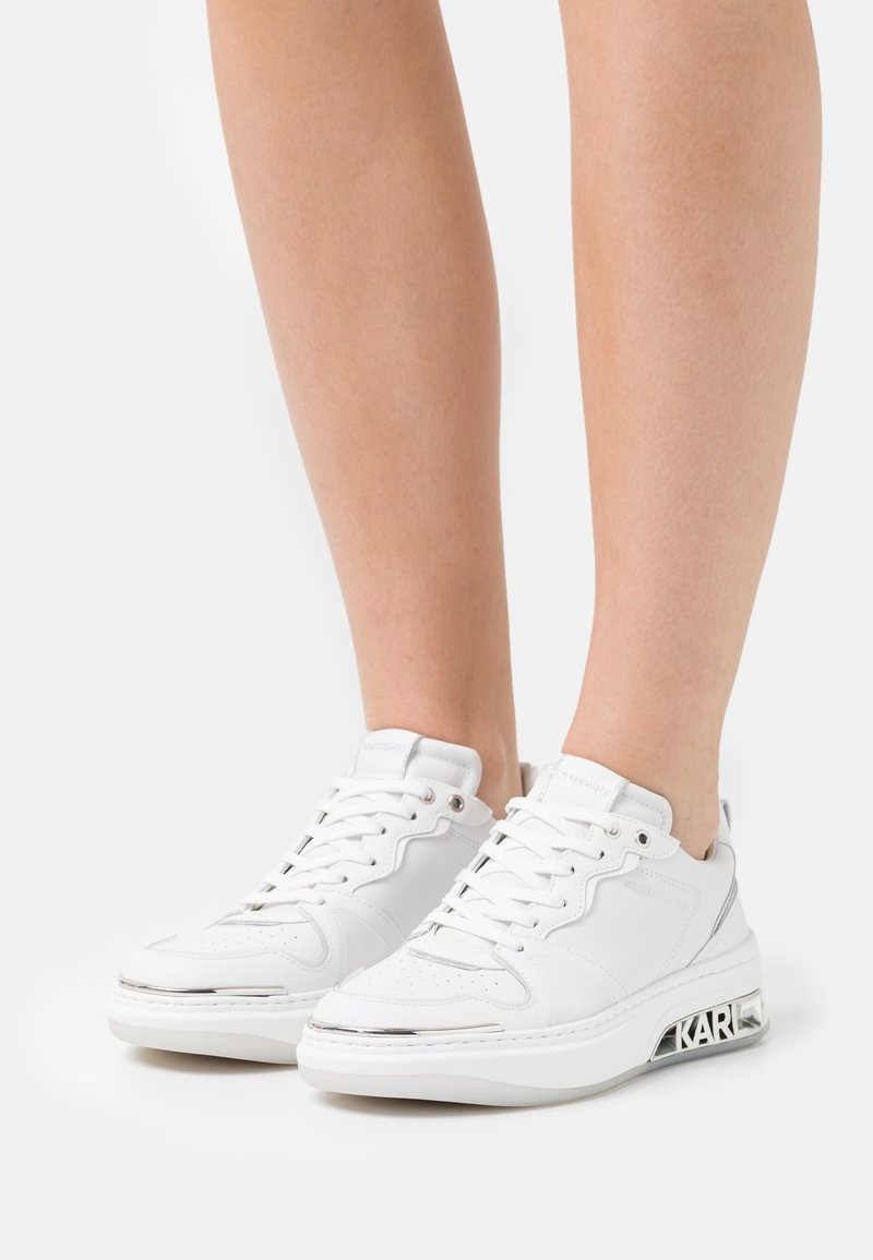 Białe sneakersy na sznurówki z grubymi podeszwami z brandingiem "KARL", noszone przez osobę stojącą na białym tle.