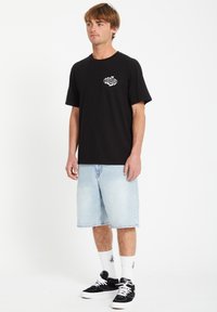 Schwarzes Baumwoll-T-Shirt mit einem weißen runden Logo, kombiniert mit hellblauen Denim-Shorts, schwarzen Sneakers und weißen gemusterten Crew-Socken.