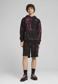 Sudadera negra con diseño de salpicaduras rosas y pantalones cortos a juego. La sudadera tiene un bolsillo canguro; el modelo lleva zapatos negros con cordones rosas.