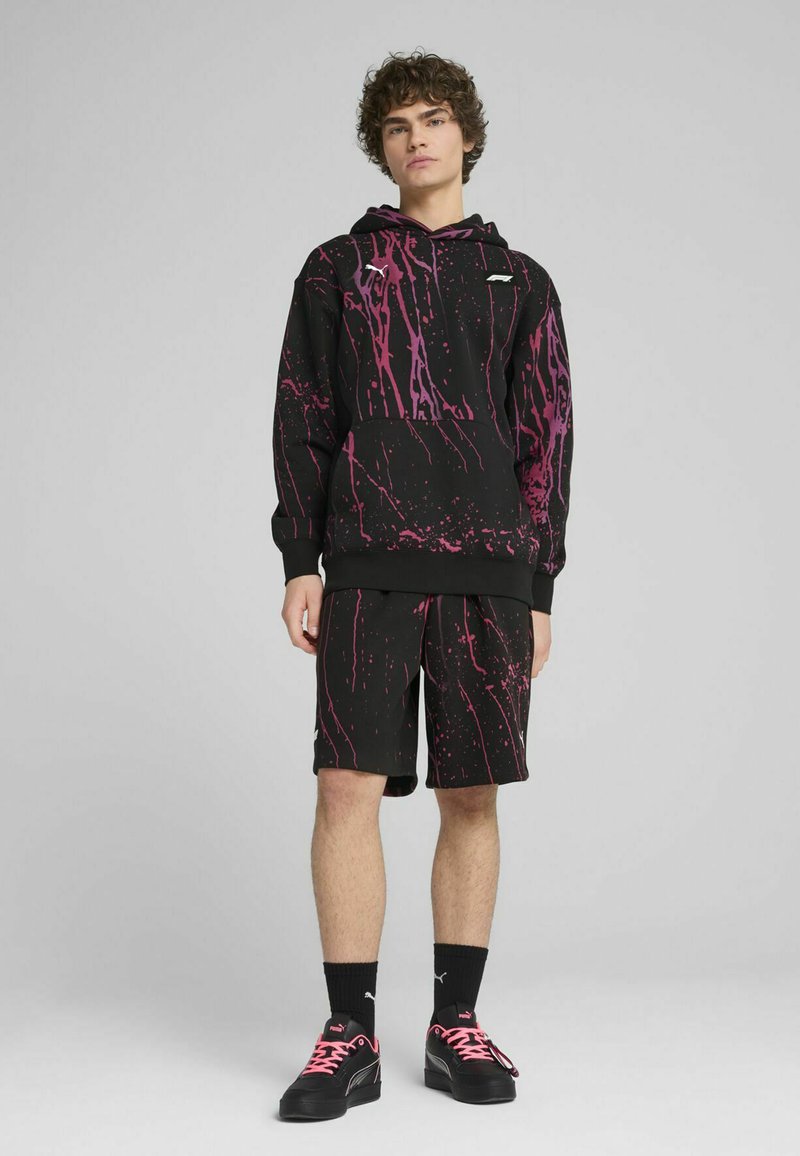 Sudadera negra con diseño de salpicaduras rosas y pantalones cortos a juego. La sudadera tiene un bolsillo canguro; el modelo lleva zapatos negros con cordones rosas.