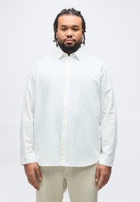 Άνδρας με πλεγμένα μαλλιά και γενειάδα, στέκεται ακίνητος φορώντας λευκό πουκάμισο με κουμπιά και μπεζ παντελόνι, μπροστά σε ένα απλό φόντο.