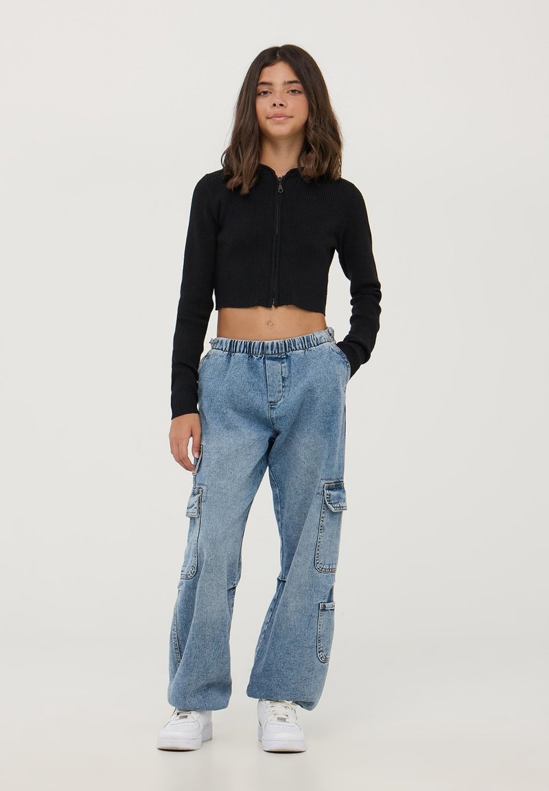 Maglione a coste neri accorciato con zip, abbinato a pantaloni cargo larghi blu chiaro con grandi tasche laterali e vita elastica. Sneakers bianche.