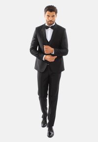 Tuxedo nero con rever in satin, camicia bianca e papillon nero; scarpe eleganti nere lucide; vestibilità su misura; postura sicura.