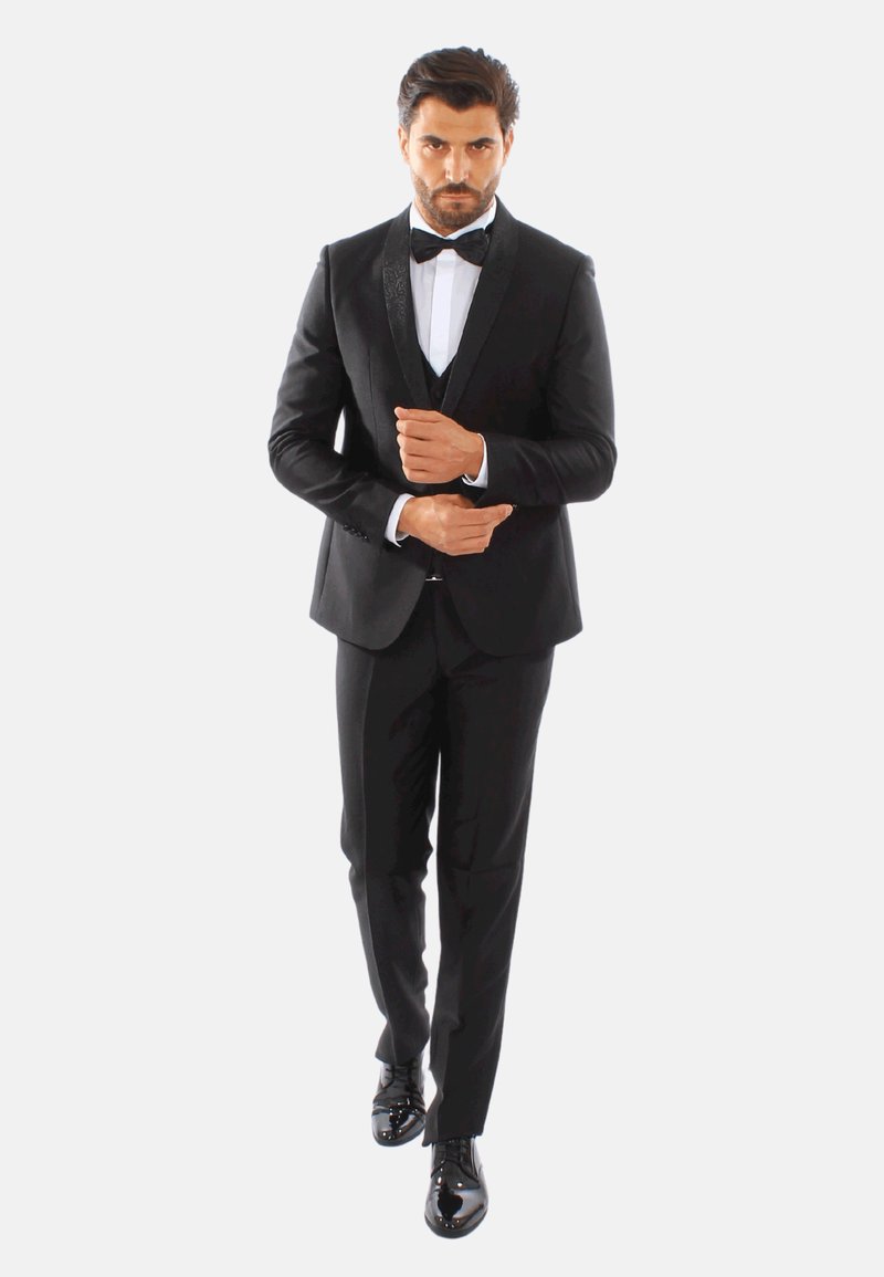 Tuxedo nero con rever in satin, camicia bianca e papillon nero; scarpe eleganti nere lucide; vestibilità su misura; postura sicura.