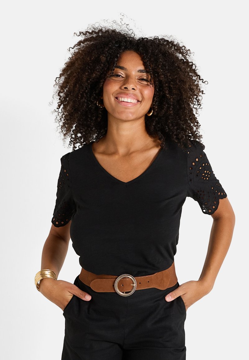 Camiseta negra de cuello en V con mangas de encaje con ojales. Combinada con un cinturón marrón con una hebilla circular de plata. Textura de tela suave.