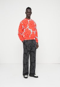 Φούτερ σε φωτεινό κόκκινο με μεγάλο λουλουδάτο μοτίβο σε ροζ και μπλε, σε συνδυασμό με μαύρο παντελόνι φαρδύ με σχέδια. Και τα δύο είναι κατασκευασμένα από απαλό ύφασμα.