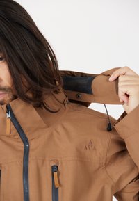 Brun vattentät jacka med hög krage, blixtlås och svart nätfoder. Funktioner inkluderar justerbara ärmar och en logotyp på axeln.