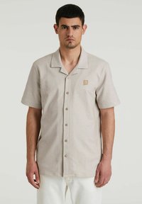 Camisa de manga corta en beige claro con rayas verticales, con un cuello tipo campamento, botones en la parte delantera y un pequeño logo en el pecho.
