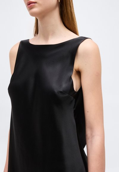 Γυναίκα που φοράει μαύρο σατέν τοπ χωρίς μανίκια με λαιμόκοψη σκάφος, εμφανιζόμενη από τους ώμους ως το μέσο του κορμού επάνω σε απλό φόντο.