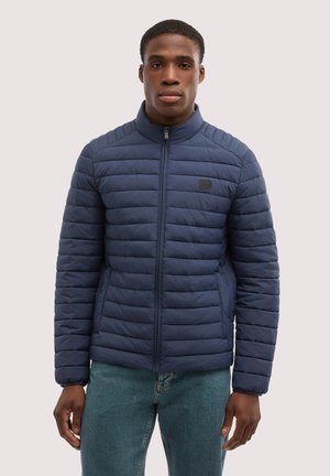 Uomo in piedi che indossa una giacca trapuntata blu navy con zip frontale e jeans azzurri chiari su uno sfondo neutro.