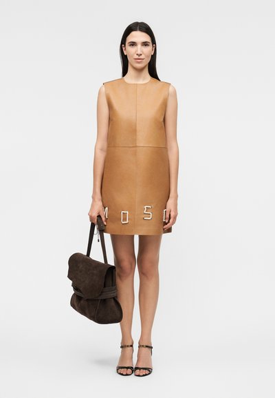 Γυναίκα με μακριά σκούρα μαλλιά φορεί μαύρο δερμάτινο φόρεμα χωρίς μανίκια με κεντημένα γράμματα "MOSO" στο τελείωμα, κρατάει σκούρα καφέ τσάντα από σουέτ και μαύρα σανδάλια.