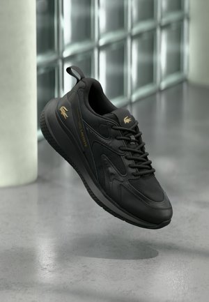 Scarpa da ginnastica nera Lacoste con logo dorato, tomaia strutturata e suola spessa sospesa sopra un pavimento in cemento grigio.