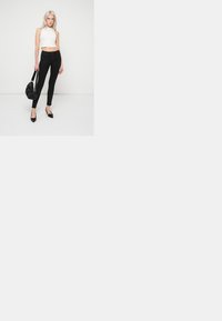 Fekete, magas derekú farmernadrág, amely testhezálló fazonú, fehér, cropped toppal párosítva. A modell egy fekete textúrált táskával a kezében és fekete magassarkú cipőben áll.