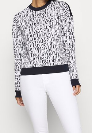 Vrouw draagt een zwart-witte trui met lange mouwen, bedrukt met herhaalde "Tommy Hilfiger" tekst en effen zwarte manchetten, gecombineerd met een witte broek.