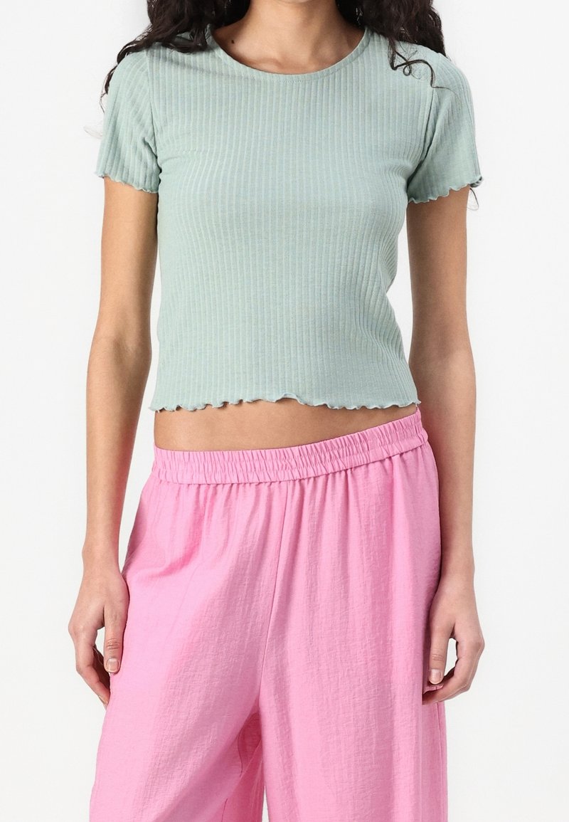 Femme portant un crop top côtelé vert clair à manches courtes et un pantalon rose ample à taille élastique, debout devant un fond blanc.