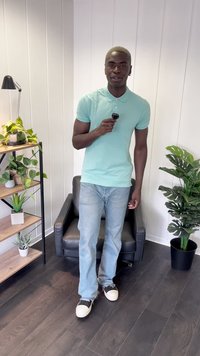 Lichtblauw kortefris polo shirt, slim-fit denim jeans en zwarte sneakers met witte zolen. Staande in een goed verlichte kamer met planten.