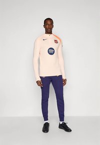 Camiseta deportiva de manga larga rosa claro con el logo azul de Spotify y el emblema rojo del FC Barcelona. Combinada con pantalones deportivos azul oscuro y zapatos negros.