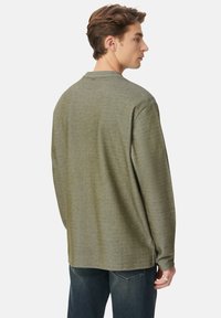 Junger Mann, der sich seitlich dreht und ein olivgrünes Langarmhemd sowie dunkelblaue Jeans trägt, vor einem einfarbigen Hintergrund.