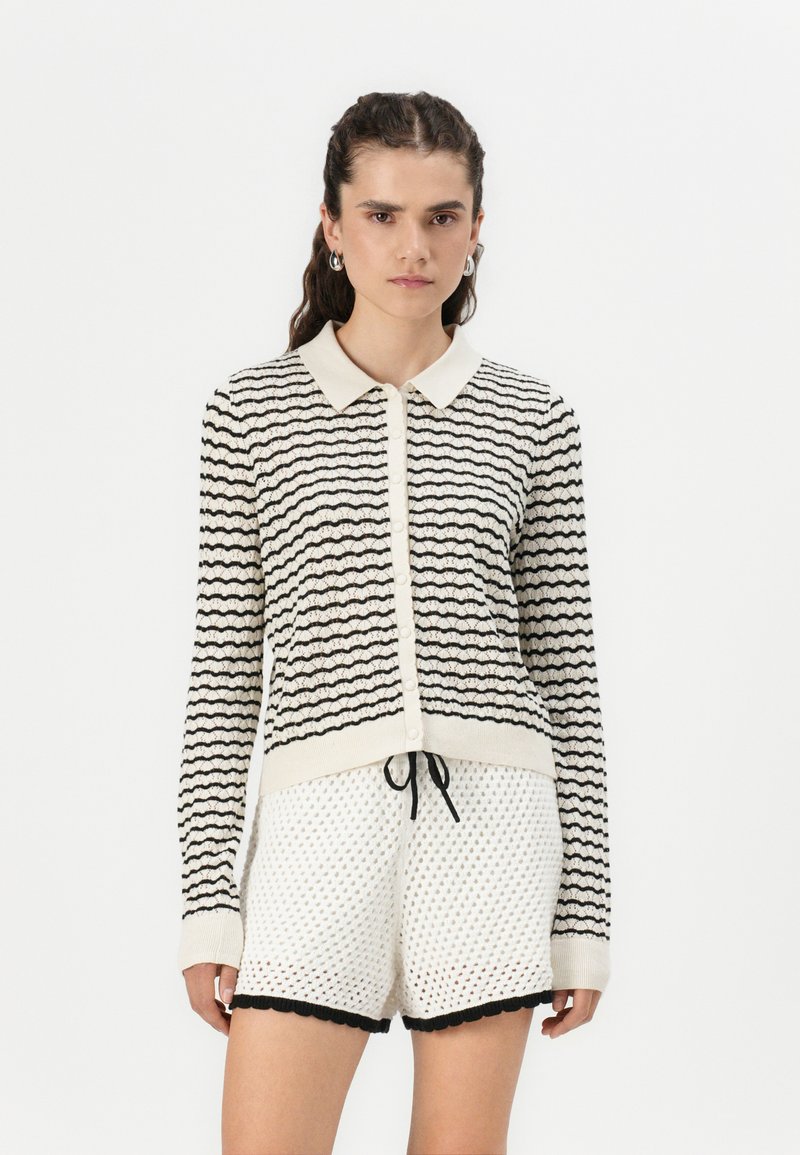 Cardigan à rayures noires et blanches avec col, tissu texturé, fermeture à boutons et design ajusté, associé à des shorts blancs à motifs.