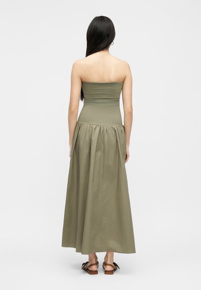 Γυναίκα με μακριά σκούρα μαλλιά, φορώντας μακρύ φόρεμα χωρίς τιράντες σε ελιά πράσινο χρώμα και καφέ σανδάλια, με την πλάτη γυρισμένη προς ένα απλό φωτεινό φόντο.