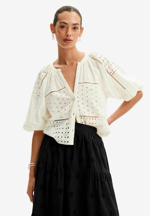 Blusa bianca ricamata con maniche corte a sbuffo, caratterizzata da motivi a occhio di pernice e una profonda scollatura a V, abbinata a una gonna nera a strati.