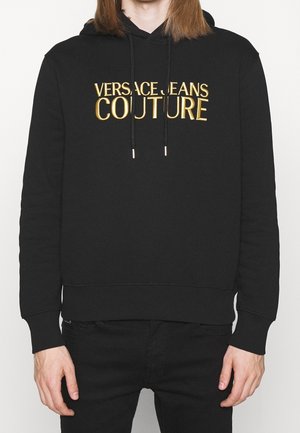 Osoba nosząca czarną bluzę z kapturem z złotym napisem "Versace Jeans Couture" na piersi, w połączeniu z czarnymi spodniami na jednolitym tle.