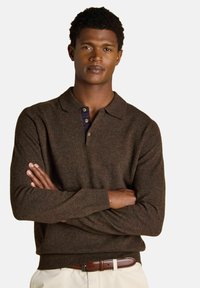 Man with folded arms wearing a brown long-sleeve polo sweater, beige pants, and a brown leather belt against a white background.
