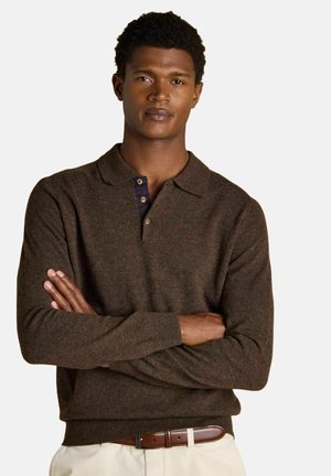 Mann mit verschränkten Armen, der einen braunen Langarm-Poloshirt, beige Hosen und einen braunen Ledergürtel vor einem weißen Hintergrund trägt.