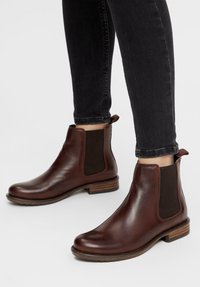 Botas de cuero marrón hasta el tobillo con paneles laterales elásticos, punteras redondas y tacones de madera apilados, combinadas con jeans ajustados negros.